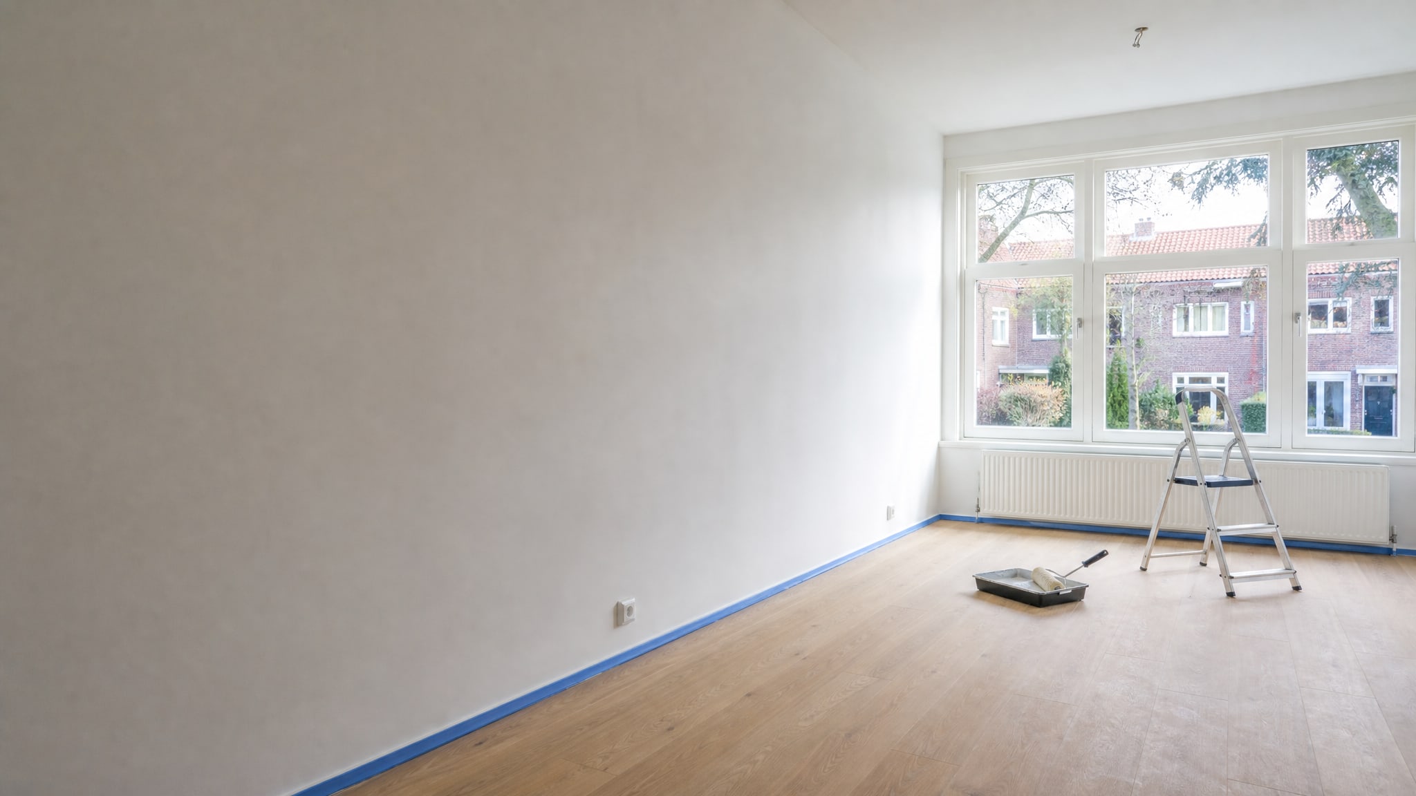Lichte woonkamer die net geschilderd is, met afplaktape langs de plint, een verfroller in de bak en een trapje bij het raam